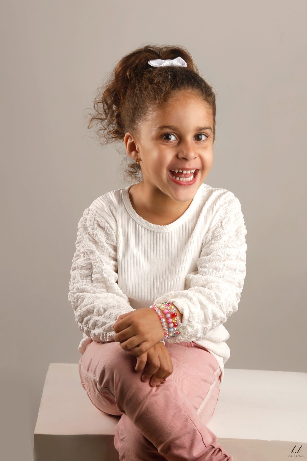 Elô sentada e sorrindo, com blusa branca e calça rosa